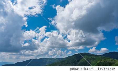 Many white clouds drift across the bright blue summer sky, and the mountains are covered in green. 139375256