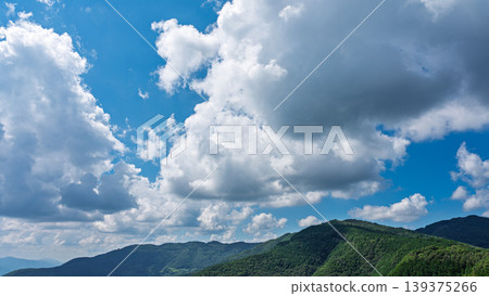 Many white clouds drift across the bright blue summer sky, and the mountains are covered in green. 139375266