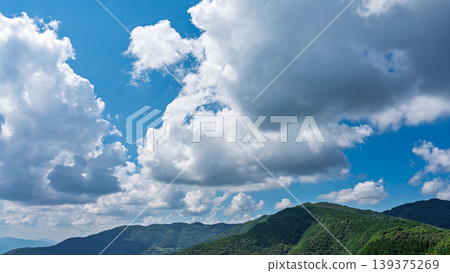Many white clouds drift across the bright blue summer sky, and the mountains are covered in green. 139375269