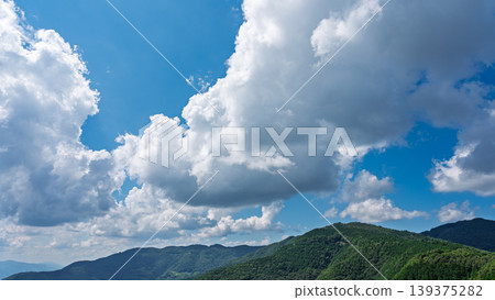 Many white clouds drift across the bright blue summer sky, and the mountains are covered in green. Many white clouds drift across the bright blue summer sky, and the mountains are covered in green. 139375282
