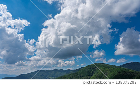 Many white clouds drift across the bright blue summer sky, and the mountains are covered in green. 139375292