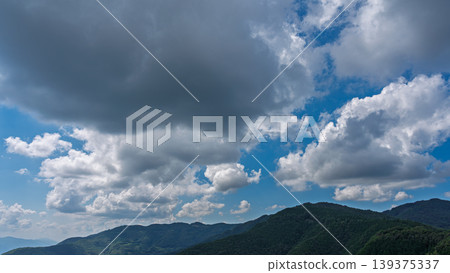 Many white clouds drift across the bright blue summer sky, and the mountains are covered in green. Many white clouds drift across the bright blue summer sky, and the mountains are covered in green. 139375337
