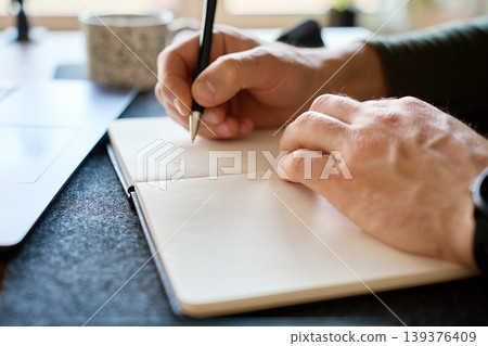 Man writing notes in notebook beside laptop at desk Man writing notes in notebook beside laptop at desk 139376409