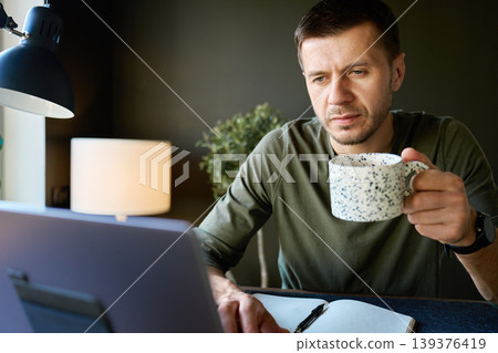Man drinking coffee while working on laptop in home office Man drinking coffee while working on laptop in home office 139376419
