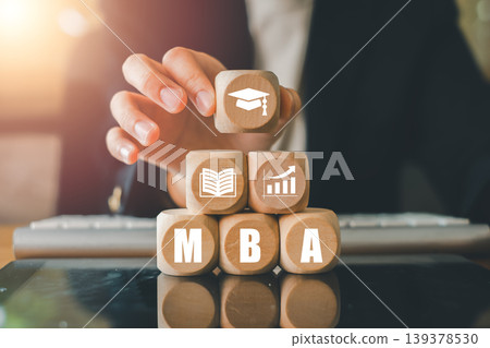 Close-up of a hand arranging wooden blocks with MBA-related icons, symbolizing education and business growth in a modern office. 139378530