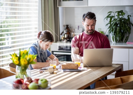 Daughter studying at table as dad works remotely on laptop nearby. 139379211