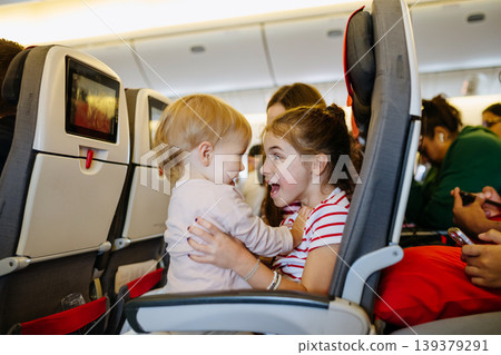 Older sister entertaining toddler sibling while sitting together on airplane seat. 139379291