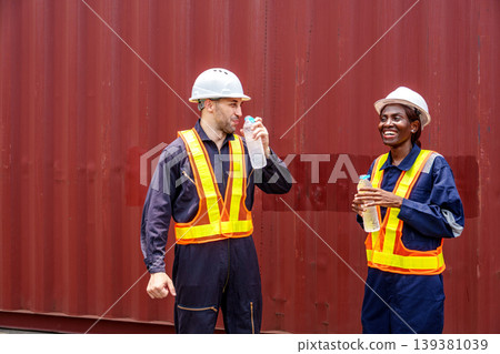 Smiling mid-adult Caucasian male and Black female industrial workers sharing a laugh while drinking water at an Asian container yard. 139381039