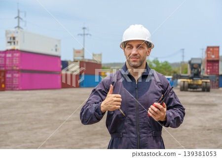 Mid-adult Caucasian male logistics worker in navy coveralls giving thumbs up at Asian shipping port container yard. Mid-adult Caucasian male logistics worker in navy coveralls giving thumbs up at Asian shipping port container yard. 139381053