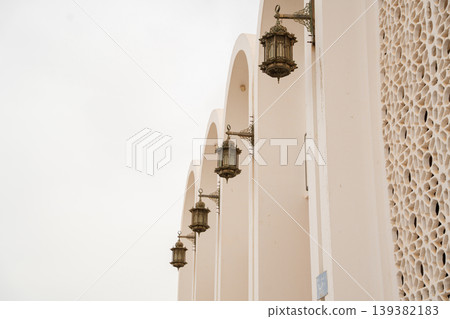 Exterior mosque wall with arched colonnade and hanging metal lanterns mounted on brackets alongside mashrabiya screen. Islamic architecture facade lighting element Exterior mosque wall with arched colonnade and hanging metal lanterns mounted on brackets alongside mashrabiya screen. Islamic architecture facade lighting element 139382183