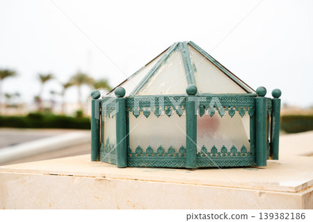 Decorative outdoor mosque lantern with frosted glass panels and metal frame placed on stone base near courtyard. Islamic architecture lighting element, exterior illumination fixture Decorative outdoor mosque lantern with frosted glass panels and metal frame placed on stone base near courtyard. Islamic architecture lighting element, exterior illumination fixture 139382186