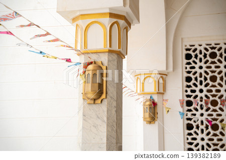 Mosque column with marble shaft and gold painted capital featuring attached wall lantern sconce and decorative mashrabiya screen in background. Islamic architecture, exterior detail, lighting 139382189