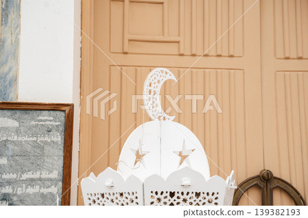 Decorative mosque lantern with crescent and star cutouts placed near wooden entrance door and inscription plaque on wall. Islamic lighting element exterior Decorative mosque lantern with crescent and star cutouts placed near wooden entrance door and inscription plaque on wall. Islamic lighting element exterior 139382193