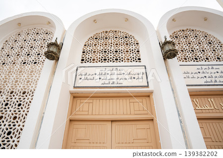 Mosque facade with arched niches mashrabiya screens wooden doors and arabic inscription panels with traditional wall lanterns. Islamic architecture exterior detail Mosque facade with arched niches mashrabiya screens wooden doors and arabic inscription panels with traditional wall lanterns. Islamic architecture exterior detail 139382202