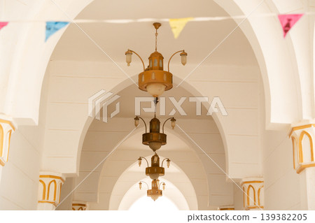 Mosque riwaq ceiling view with pointed arches and suspended lanterns aligned along central axis with decorative gold column capitals. Islamic architecture, interior detail, structural element Mosque riwaq ceiling view with pointed arches and suspended lanterns aligned along central axis with decorative gold column capitals. Islamic architecture, interior detail, structural element 139382205