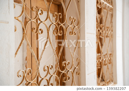 Close up of mosque window wrought iron grille with curved scroll pattern mounted over wooden shutters in white wall frame. Islamic architecture, decorative metalwork, exterior detail 139382207