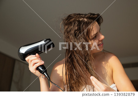 Beauty woman holding hairdryer and drying long wet hair, indoor portrait with soft sunlight, female model with smooth skin and relaxed expression, haircare and lifestyle concept 139382879
