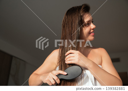 Beautiful woman brushing long wet hair with black brush in warm sunlight, beauty haircare routine at home, female model with smooth skin and relaxed pose, wellness lifestyle and daily selfcare concept 139382881
