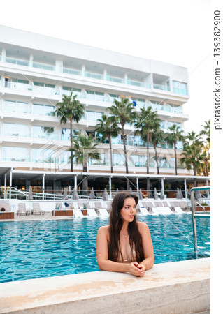 Beautiful woman leaning on pool edge near luxury resort building with palm trees enjoying summer vacation travel lifestyle and beauty relaxation under sunlight Beautiful woman leaning on pool edge near luxury resort building with palm trees enjoying summer vacation travel lifestyle and beauty relaxation under sunlight 139382900