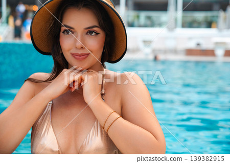 Beauty woman posing near pool with straw hat touching chin enjoying summer vacation luxury resort lifestyle portrait with glowing skin and relaxed expression 139382915