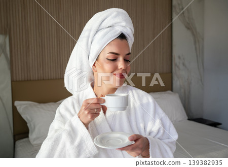 Woman in white robe and towel enjoys coffee in hotel room after morning routine before starting the day 139382935