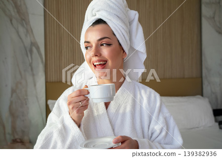 Smiling woman in robe holding cup of coffee while relaxing in hotel room during morning hours Smiling woman in robe holding cup of coffee while relaxing in hotel room during morning hours 139382936