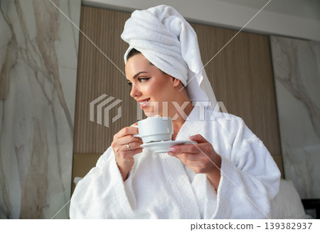 Woman in a white robe holds a cup of coffee in a modern room while wearing a towel on her head in the morning light after a refreshing shower 139382937