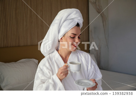 Woman enjoys coffee while wearing a robe and towel in a hotel room during morning hours Woman enjoys coffee while wearing a robe and towel in a hotel room during morning hours 139382938