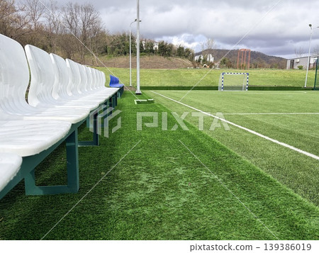 A soccer field, featuring a row of white plastic stadium seats with green metal frames running along the left edge. A soccer field, featuring a row of white plastic stadium seats with green metal frames running along the left edge. 139386019