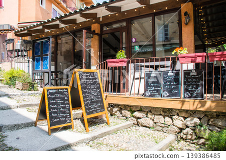 Pastel-colored buildings and chairs and tables placed in front of a restaurant on the French island of Corsica in the Mediterranean Sea. 139386485