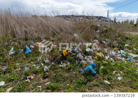 Various pieces of garbage are lying on the grass in an urban setting next to a landfill surrounded by tall grass and weeds 139387216