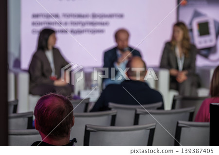 Business conference panel seen from audience, blurred speakers on stage and attentive attendees Business conference panel seen from audience, blurred speakers on stage and attentive attendees 139387405