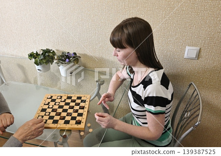 Two people play checkers at a glass table with plants in a light room during the daytime 139387525