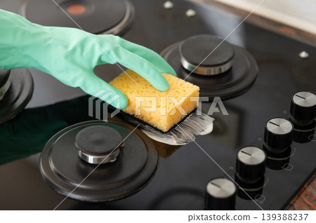 A close-up of a person's hand in a rubber glove cleaning a gas cooktop with a sponge. A close-up of a person's hand in a rubber glove cleaning a gas cooktop with a sponge. 139388237