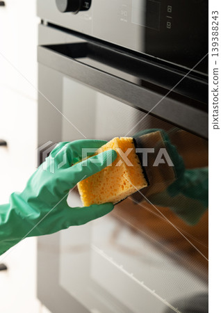 Cleaning the oven door with a sponge and a rubber glove, removing grease in a modern kitchen. Cleaning the oven door with a sponge and a rubber glove, removing grease in a modern kitchen. 139388243
