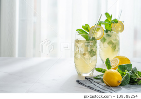 A summer drink with lemon, mint, and ice in a two glasses against a sunny window background. A summer drink with lemon, mint, and ice in a two glasses against a sunny window background. 139388254