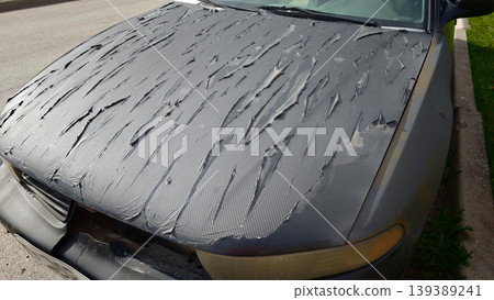 Decorative film peeled off the hood of a car revealing damaged surface underneath, with visible texture and wear in an outdoor setting Decorative film peeled off the hood of a car revealing damaged surface underneath, with visible texture and wear in an outdoor setting 139389241