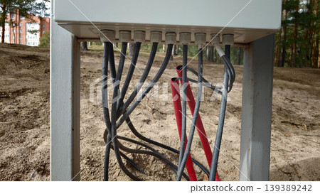 Electrical junction box with multiple cables connected, featuring gray and red wires, positioned on sandy ground in a forested area with trees in the background Electrical junction box with multiple cables connected, featuring gray and red wires, positioned on sandy ground in a forested area with trees in the background 139389242