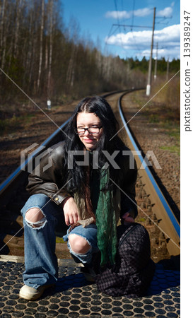 Young woman with long black hair wearing a brown leather jacket and ripped jeans squats on railway tracks surrounded by trees and blue sky in a rural setting Young woman with long black hair wearing a brown leather jacket and ripped jeans squats on railway tracks surrounded by trees and blue sky in a rural setting 139389247