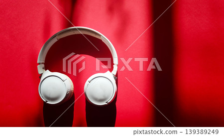 White over-ear headphones positioned on a vibrant red surface, with soft shadows creating a dynamic visual contrast and highlighting the sleek design of the headphones White over-ear headphones positioned on a vibrant red surface, with soft shadows creating a dynamic visual contrast and highlighting the sleek design of the headphones 139389249