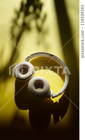 White over-ear headphones resting on a yellow surface with soft shadows and a hint of foliage in the background, showcasing a minimalist aesthetic and design 139389265