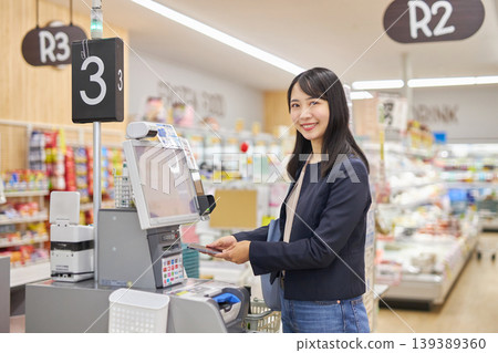 一位年輕女子正在使用智慧型手機在自助結帳櫃檯付款。 139389360