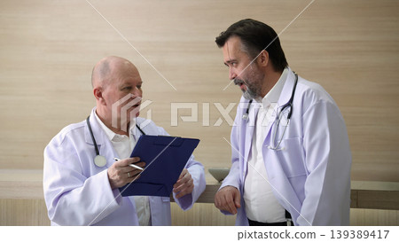 Doctors discuss. Two experienced doctors discussing patient information and medical reports in a hospital corridor. Professional healthcare teamwork consultation and collaboration in clinic hospital 139389417