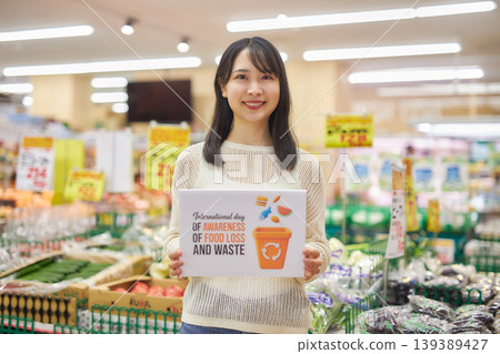 A young woman holding a sign about food waste in a supermarket aisle. A young woman holding a sign about food waste in a supermarket aisle. 139389427