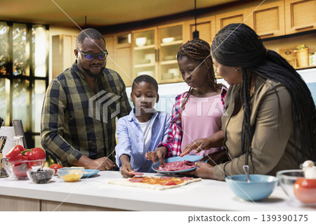 Black young kids and their parents cooking homemade pizza together, slicing fresh ingredients, sprinkling ingredients and sharing laughter at the kitchen island counter. Family activity. Black young kids and their parents cooking homemade pizza together, slicing fresh ingredients, sprinkling ingredients and sharing laughter at the kitchen island counter. Family activity. 139390175