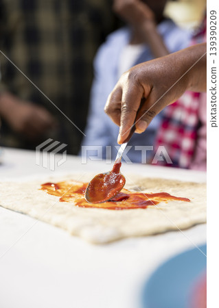 Close up of african american parents and kids using tomato sauce on the dough, preparing a homemade pizza with fresh ingredients before baking. Family fun and bonding time at home. Close up of african american parents and kids using tomato sauce on the dough, preparing a homemade pizza with fresh ingredients before baking. Family fun and bonding time at home. 139390209
