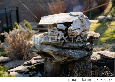 Stack of flat stones arranged as small garden structure on wooden stump outdoors in sunlight. Landscaping and natural decoration Stack of flat stones arranged as small garden structure on wooden stump outdoors in sunlight. Landscaping and natural decoration 139390257