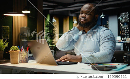 African american male worker stretching his back and neck muscles, correcting posture for muscle relief and relaxation. Strain and neck stiffness due to intense activity in office. African american male worker stretching his back and neck muscles, correcting posture for muscle relief and relaxation. Strain and neck stiffness due to intense activity in office. 139390311