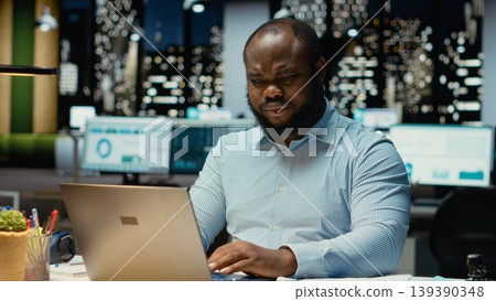 African american man does critical thinking and proofreading with papers before transcribing data on notebook, sitting in the office at night. Worker documenting important ideas. African american man does critical thinking and proofreading with papers before transcribing data on notebook, sitting in the office at night. Worker documenting important ideas. 139390348