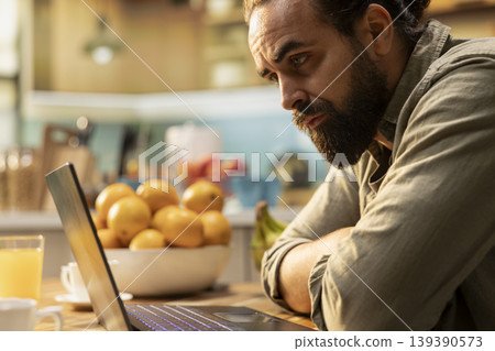 Male frowning, laptop screen, disoriented and unsure, uncertain disorganized man, error or misunderstanding at kitchen table. Ambiguous confused freelancer, preoccupied telecommuting. 139390573